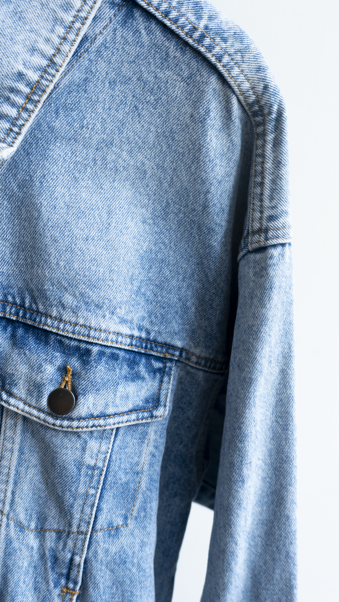 Close-up of a vintage 90s light blue denim jacket showing the chest pocket with a dark metal snap button and the textured denim fabric